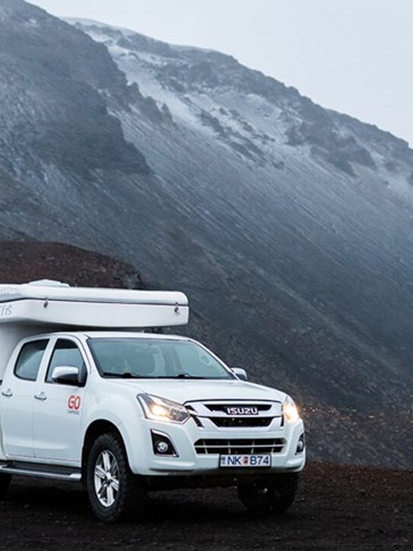 White camper truck on dark, rugged terrain with a snow-capped mountain in the background.
