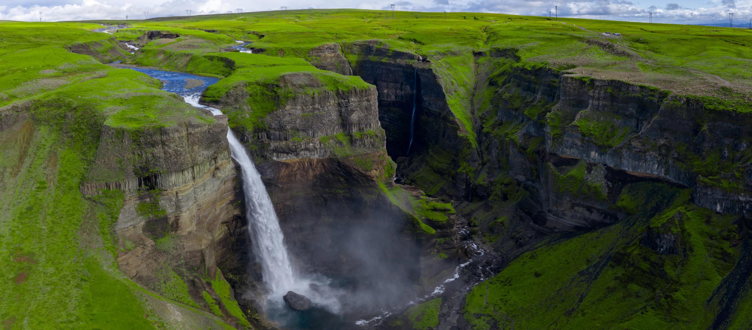 A large waterfall cascades into a deep, vibrant green canyon, with a smaller waterfall visible in the background.