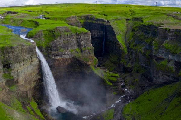 A large waterfall cascades into a deep, vibrant green canyon, with a smaller waterfall visible in the background.
