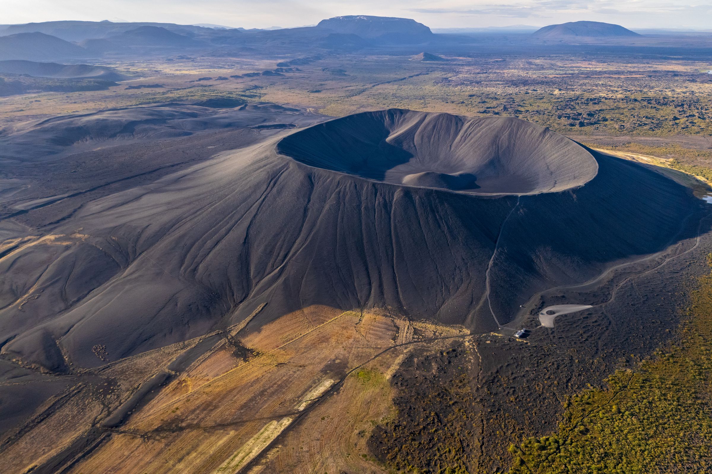 Vista aérea de un gran cráter volcánico oscuro, rodeado de un paisaje escarpado con roca volcánica negra, tierra marrón y vegetación verde.