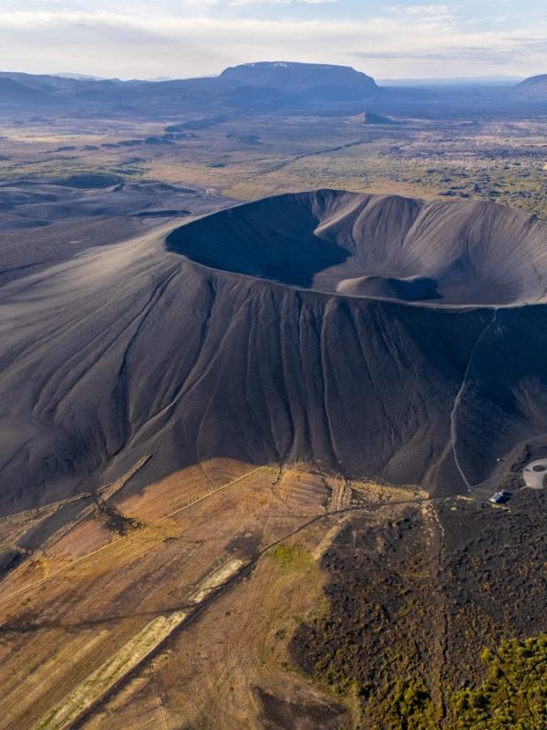 Aerial view of a dark, wide volcanic crater surrounded by barren, rugged terrain and distant hills.
