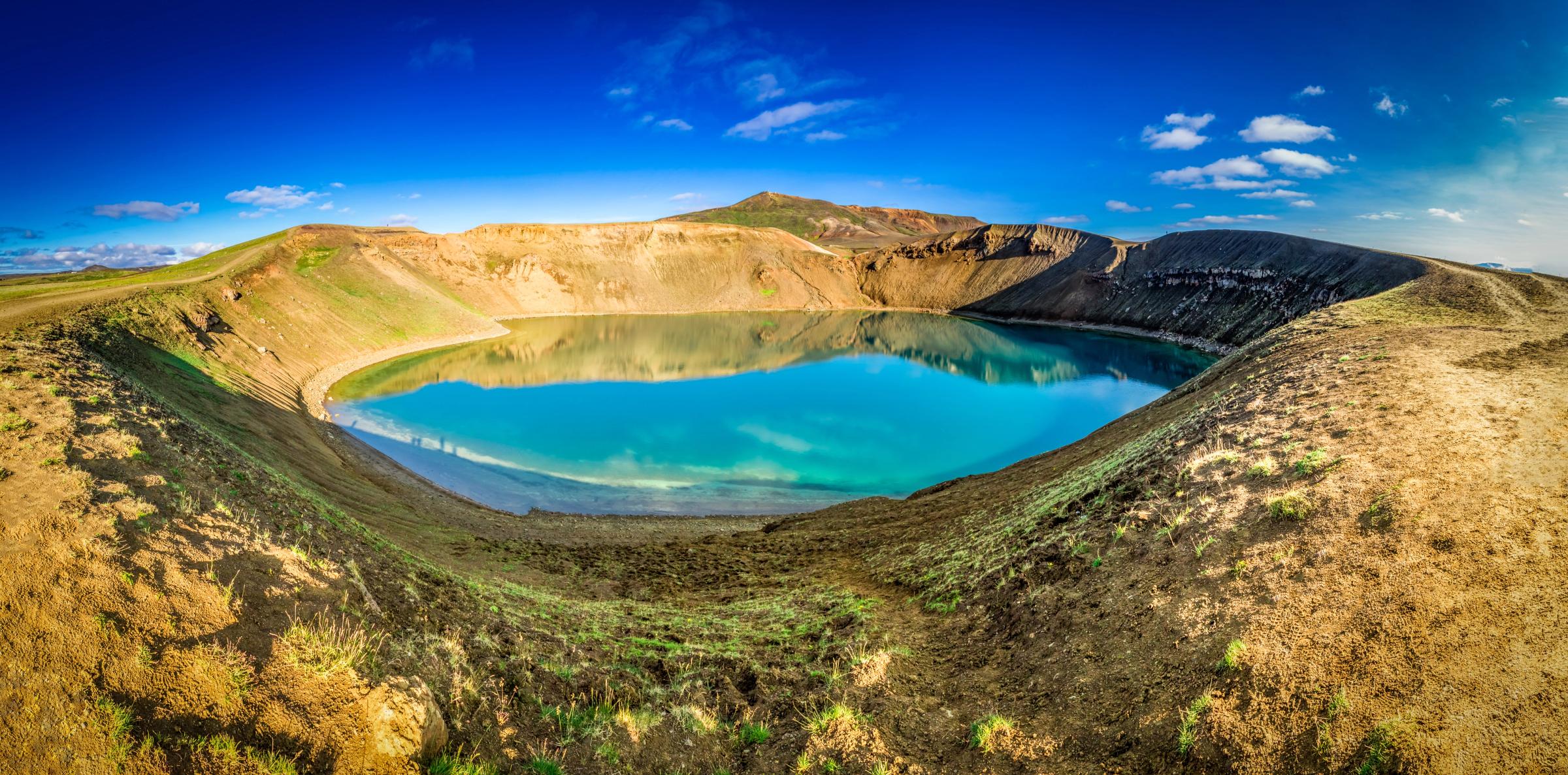 Panoramic view of a turquoise crater lake surrounded by brown and green slopes under a bright blue sky.