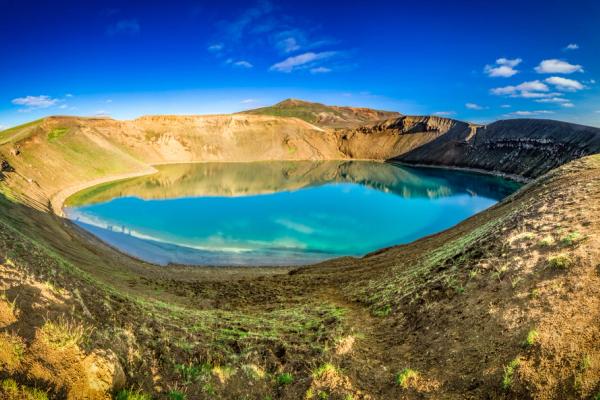 Panoramic view of a turquoise crater lake surrounded by brown and green slopes under a bright blue sky.