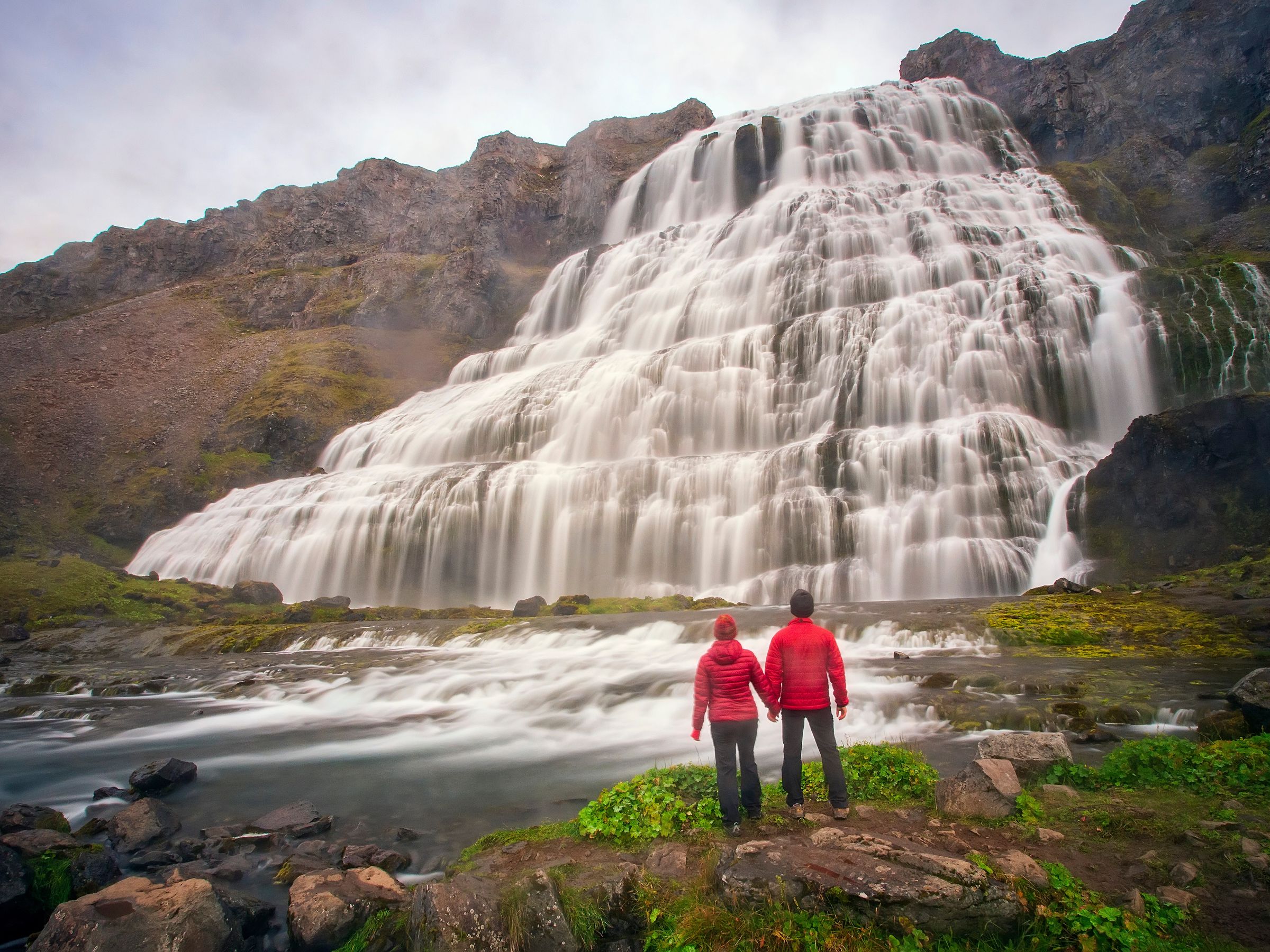 a man and a woman are standing in front of Dynjandi waterfall .