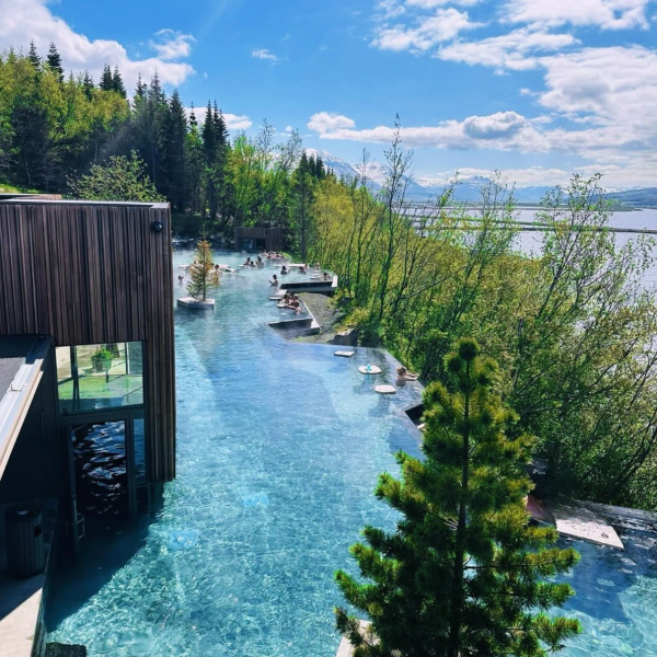Outdoor thermal lagoon with people relaxing, surrounded by trees, overlooking a lake and distant mountains.