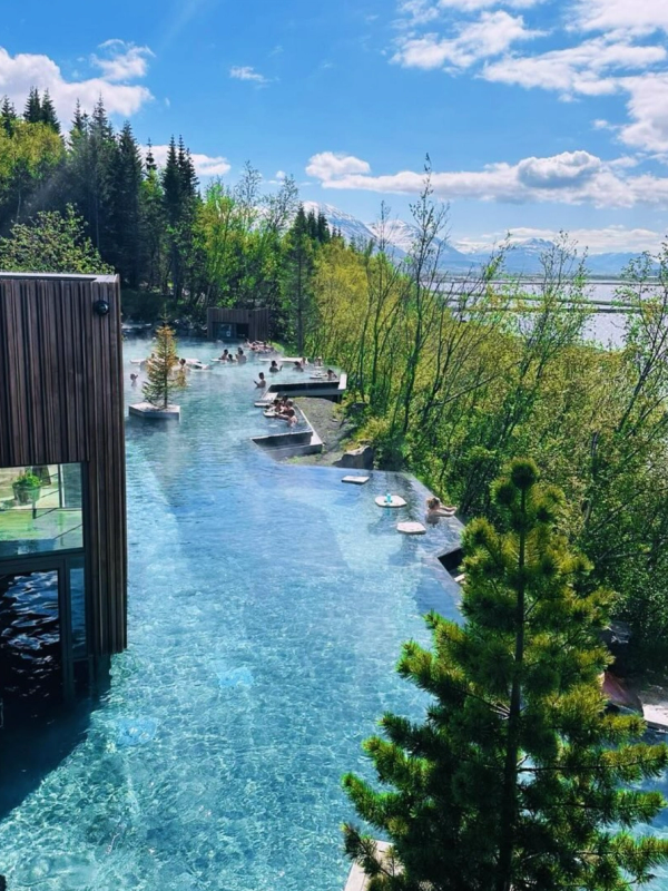 An outdoor geothermal spa with blue pools and people relaxing, surrounded by lush trees, overlooking a lake and mountains on a sunny day.