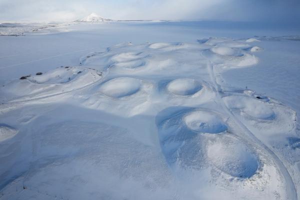 Vista aérea de un campo de pseudocráteres cubierto de nieve, con un lago congelado y una montaña distante.