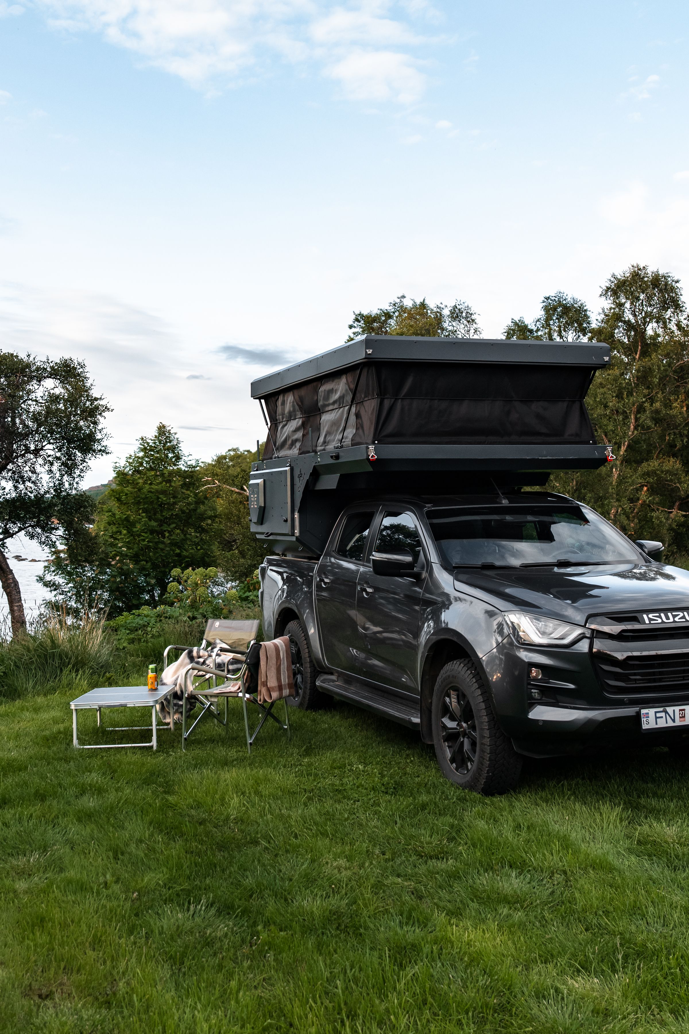 Montaje del campamento con techo elevable Cámper con techo elevable levantado junto a unas sillas y una mesa sobre el césped, alquiler de furgoneta cámper 4x4 en Islandia