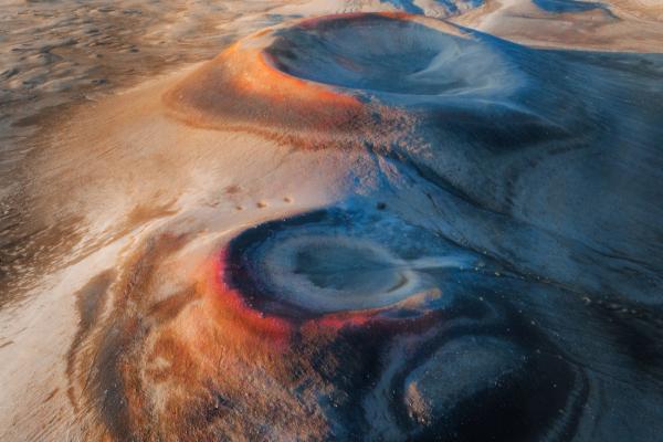 Aerial view of vibrant desert formations in shades of orange, red, and blue under a cloudy sky.