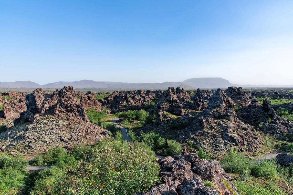 Un paisaje volcánico lleno de formaciones rocosas oscuras y escarpadas, arbustos verdes, un sendero serpenteante y montañas lejanas bajo un cielo despejado.