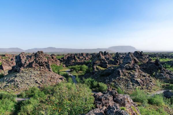 A volcanic landscape filled with dark, jagged rock formations, green shrubs, a winding path, and distant mountains under a clear sky.