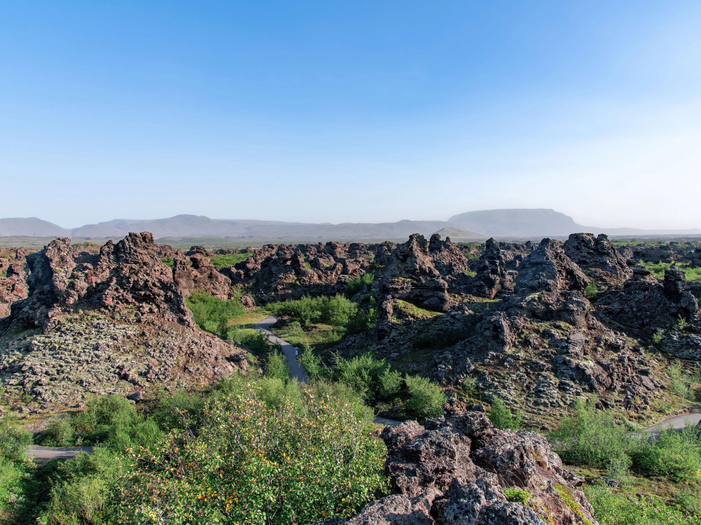 A rugged landscape of dark volcanic rock formations and green vegetation under a clear blue sky.