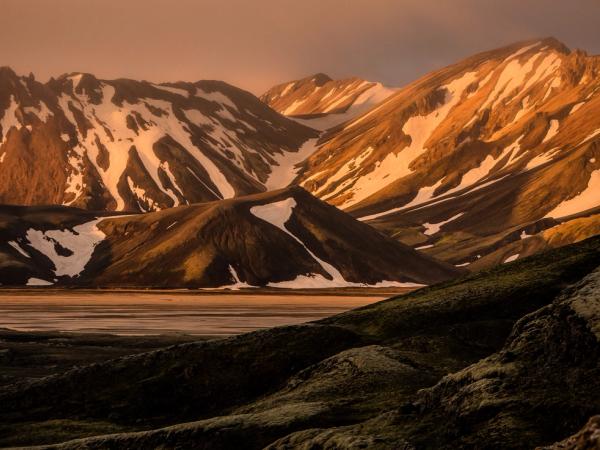 a mountain range with snow on the peaks and a lake in the foreground in iceland mightnight sun.