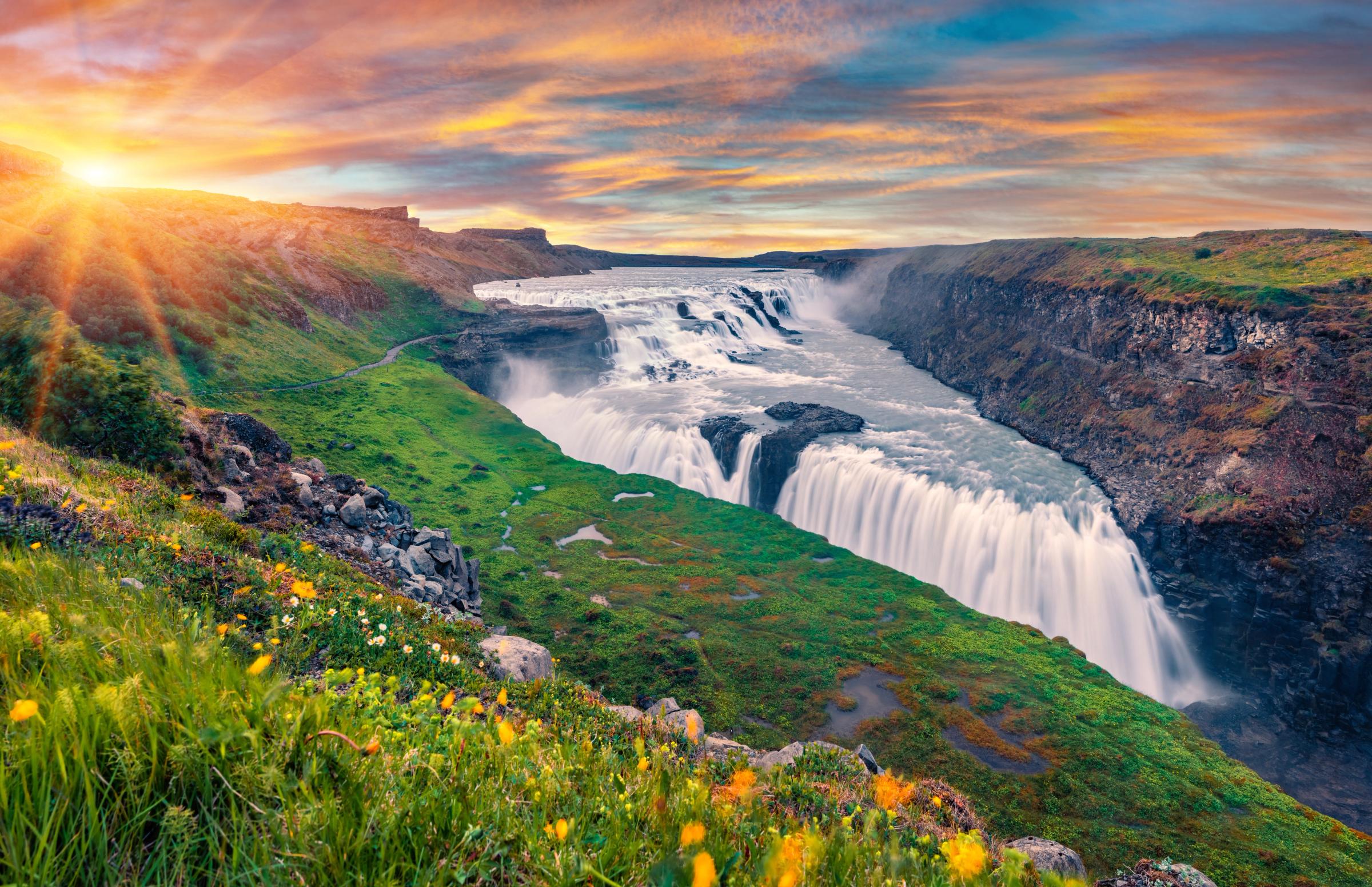 una cascada en medio de un valle con flores en primer plano y el sol brillando a través de las nubes.