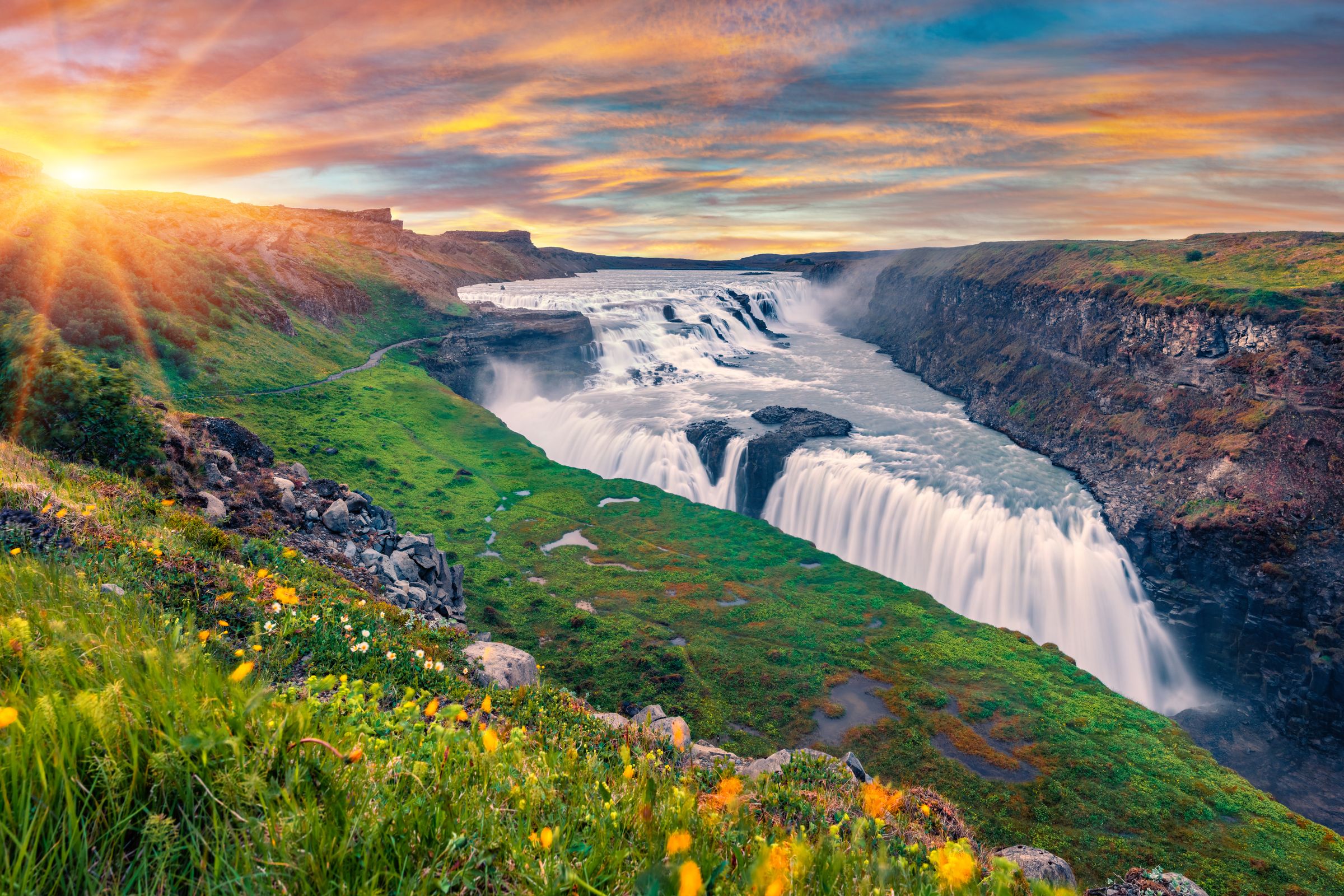 una cascada en medio de un valle con flores en primer plano y el sol brillando a través de las nubes.