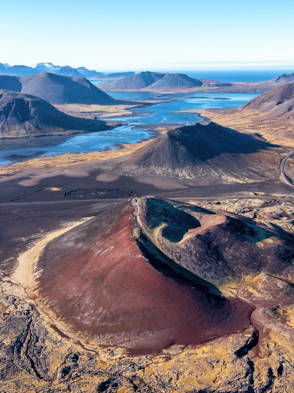 Aerial view of a volcanic landscape with a prominent reddish cinder cone, dark lava fields, a blue fjord, and distant mountains under a clear sky.