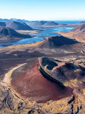 Aerial view of a volcanic landscape with a prominent reddish cinder cone, dark lava fields, a blue fjord, and distant mountains under a clear sky.