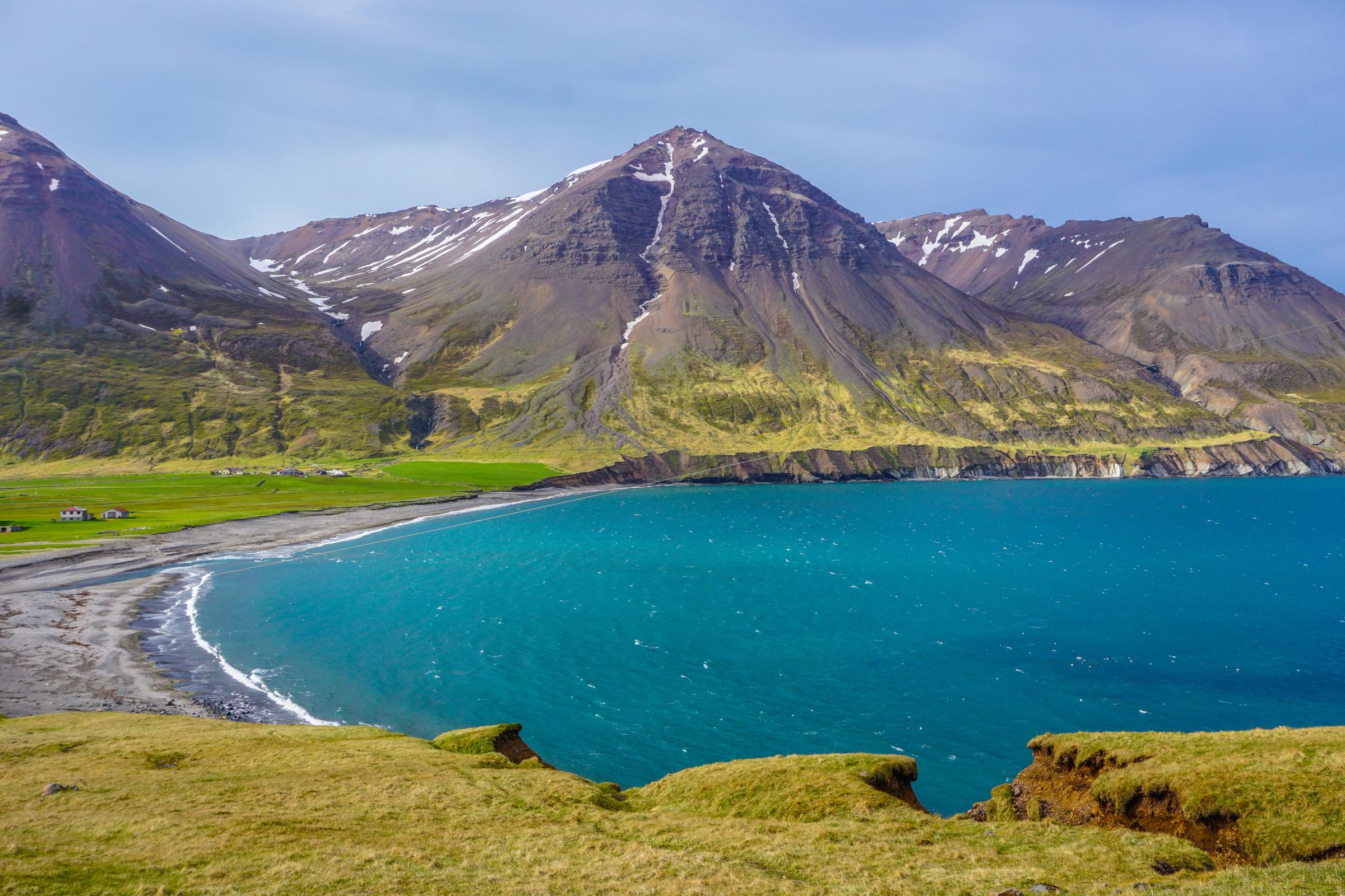 Una bahía de un turquesa vibrante con una playa de arena oscura, rodeada de colinas verdes y montañas nevadas.