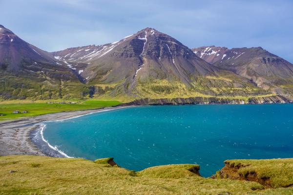 Una bahía de un turquesa vibrante con una playa de arena oscura, rodeada de colinas verdes y montañas nevadas.