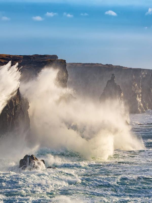 A bright orange lighthouse on a cliff with large waves crashing against the base.