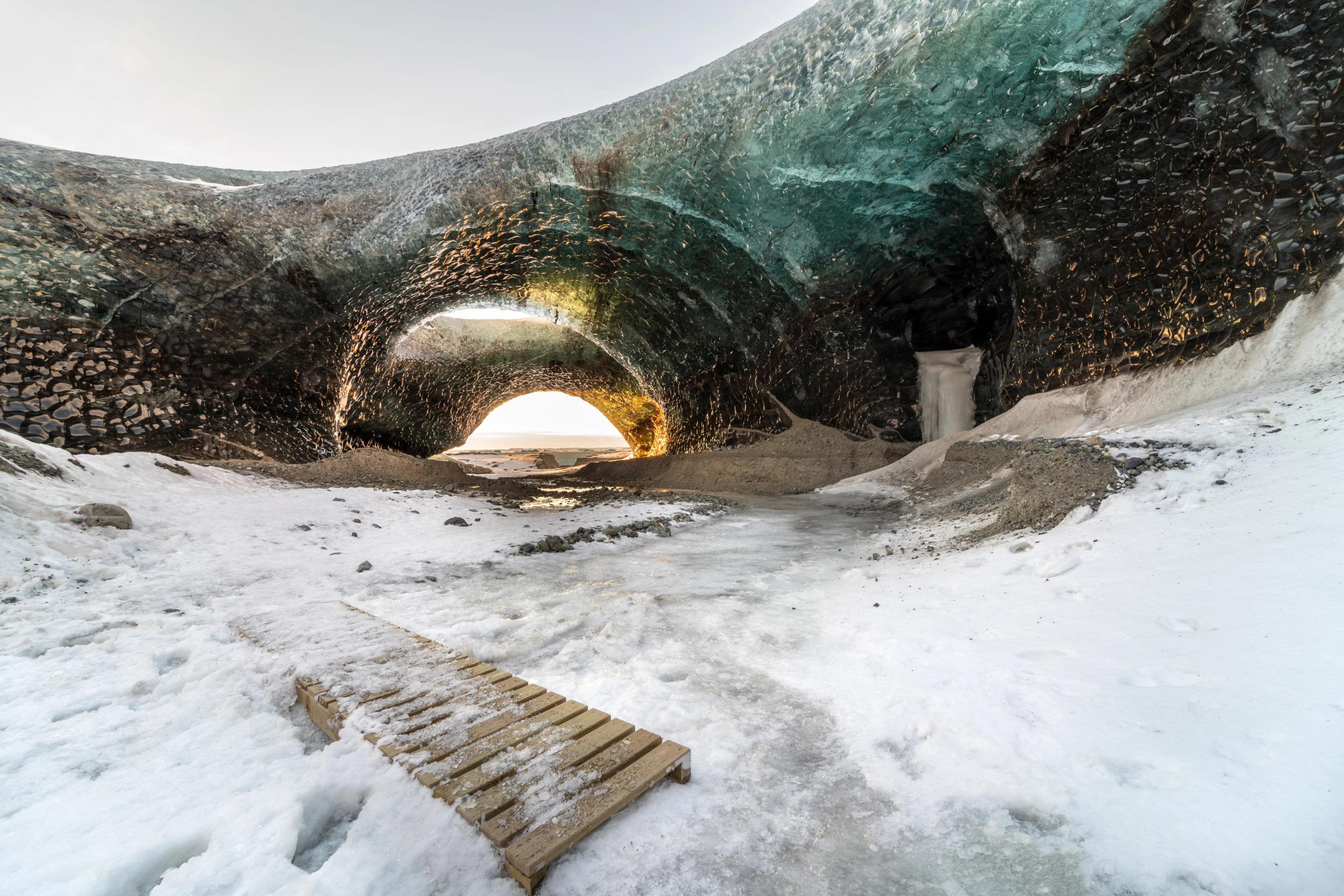 Interior de una cueva de hielo con una abertura en forma de arco por la que entra una luz intensa, suelo nevado y una tabla de madera.
