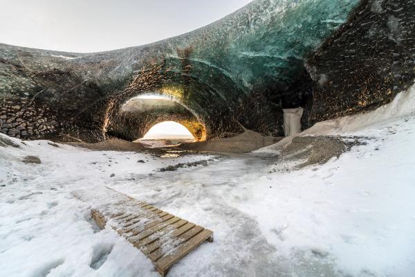 Ice cave interior with an arched opening showing bright light, snowy ground, and a wooden plank.