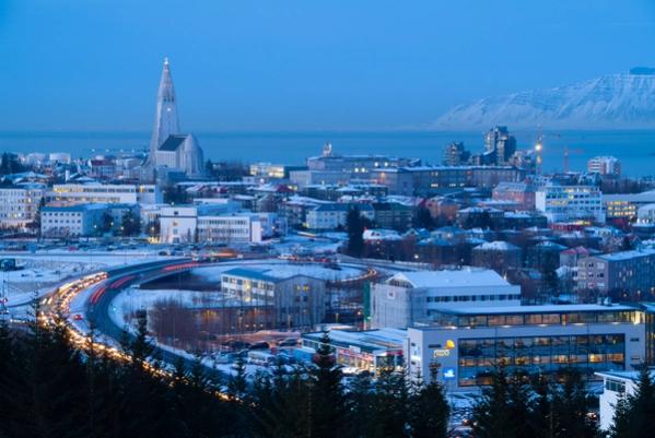 Twilight view of a snow-covered city featuring Hallgrímskirkja church, light trails from traffic, and distant mountains.