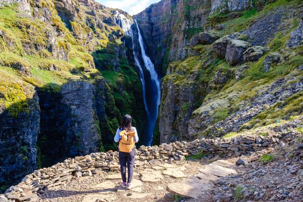 A person with a yellow backpack stands on a rocky path, looking at a tall waterfall in a green canyon.