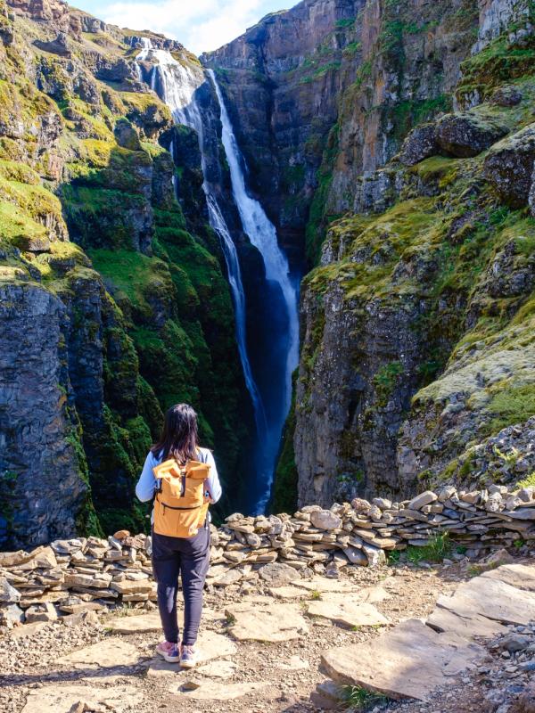 A hiker with a yellow backpack stands on a rocky path looking at a tall waterfall in a green canyon.