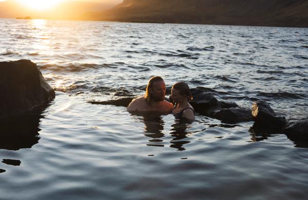 A man and a woman relaxing in a natural pool with a sunset over a lake and mountains.