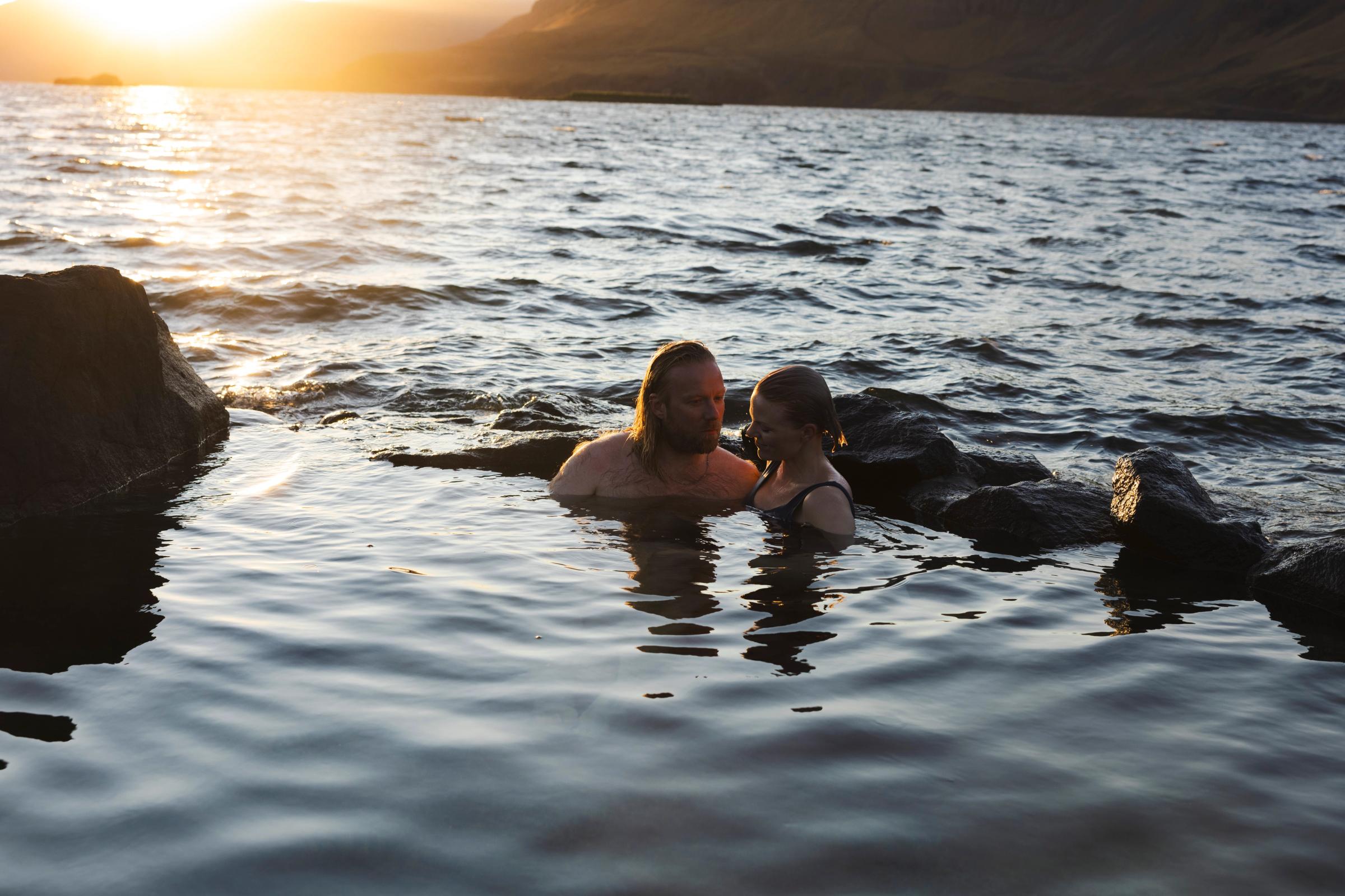 A man and a woman in a rocky natural pool at sunset with mountains in the background.
