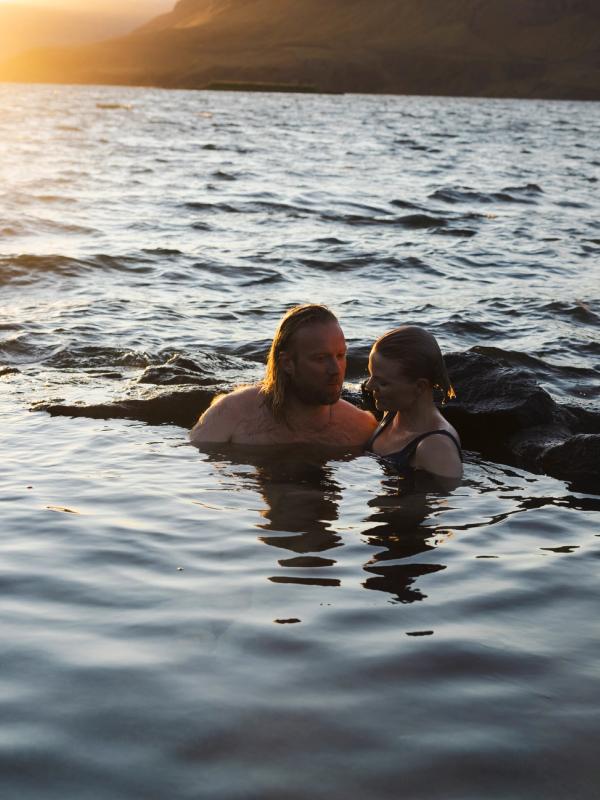 A man and a woman relaxing in a natural pool with a sunset over a lake and mountains.