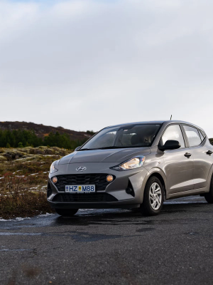 A light gray Hyundai i10 hatchback parked on a country road with a mossy, rocky landscape and evergreen trees.