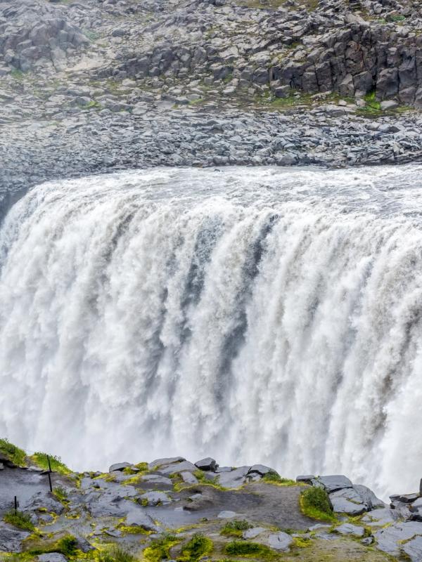 A wide waterfall plunges into a rocky canyon, with people on a viewing path.