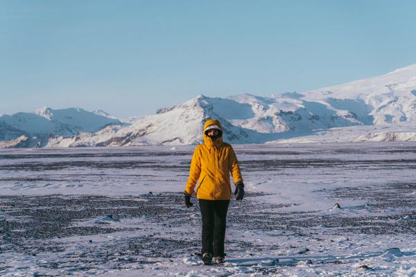 Una persona con una chaqueta amarilla está de pie en una llanura negra cubierta de nieve, con montañas nevadas bajo un cielo azul.