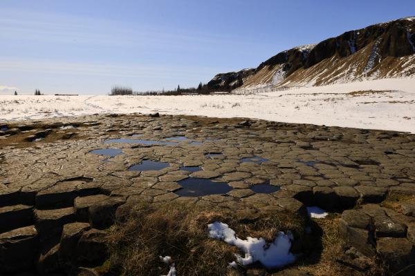Hexagonal basalt columns with puddles in the foreground, next to a snow-covered field and a mountain under a clear blue sky.