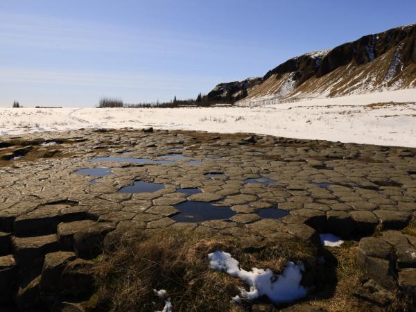Hexagonal basalt columns with water puddles in a snowy landscape with hills under a blue sky.