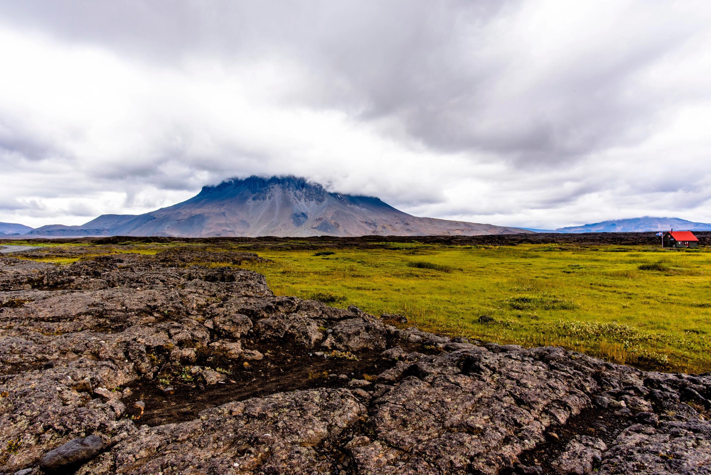 a red house is sitting in the middle of a grassy field with a mountain in the background .