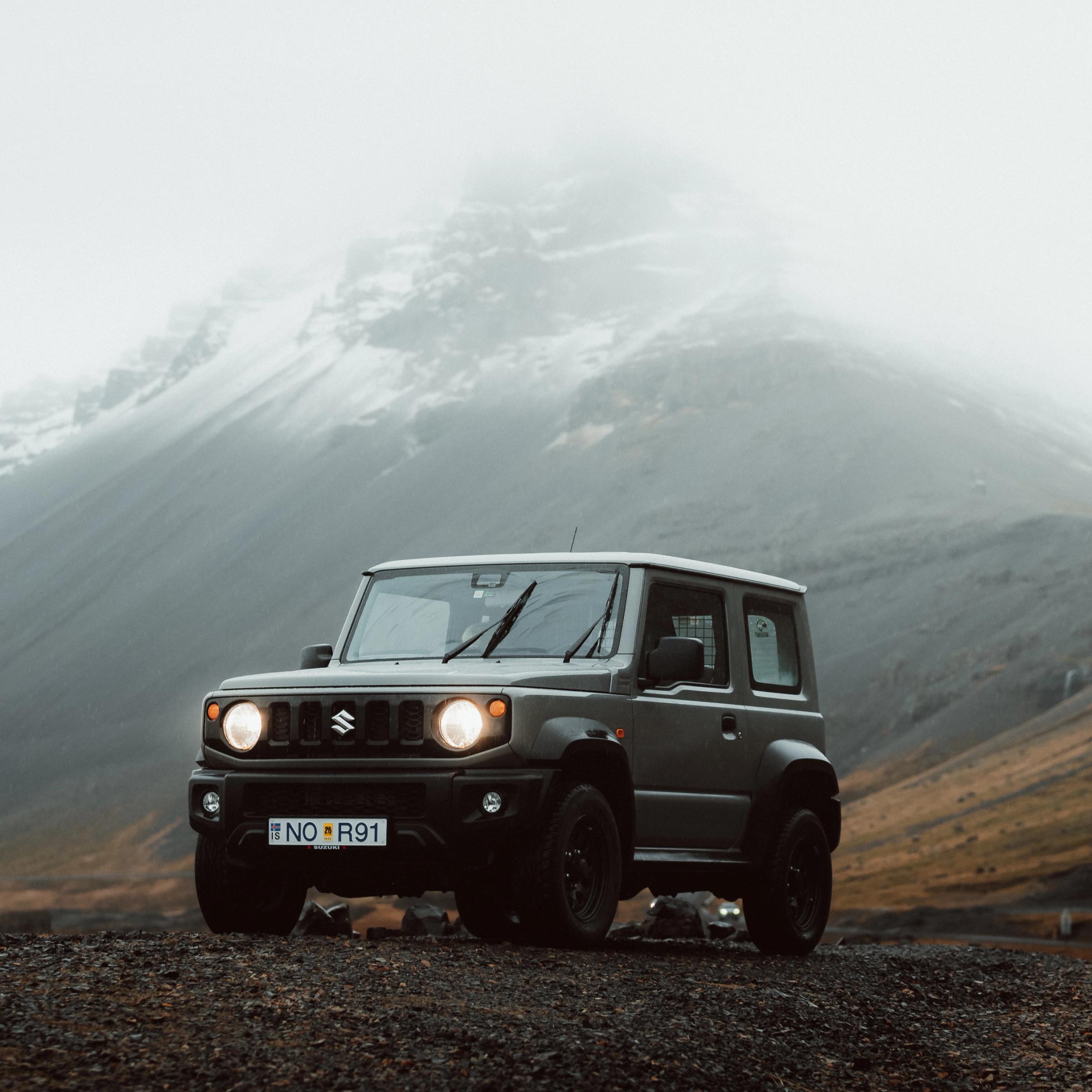 SUV Suzuki Jimny gris oscuro en una carretera de grava, con montañas nevadas y cubiertas de niebla al fondo.