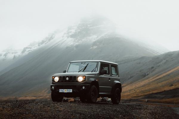 A grey Suzuki Jimny SUV with headlights on, parked on rocky ground before misty, snow-capped mountains.