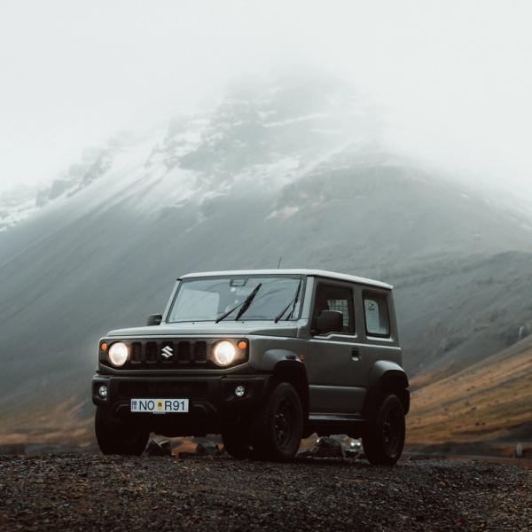 a suzuki jimny is parked on a dirt road in front of a mountain .