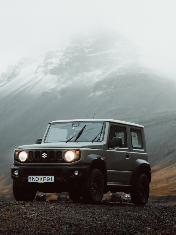 Dark green Suzuki Jimny parked on a gravel road with misty, snow-capped mountains in the background.
