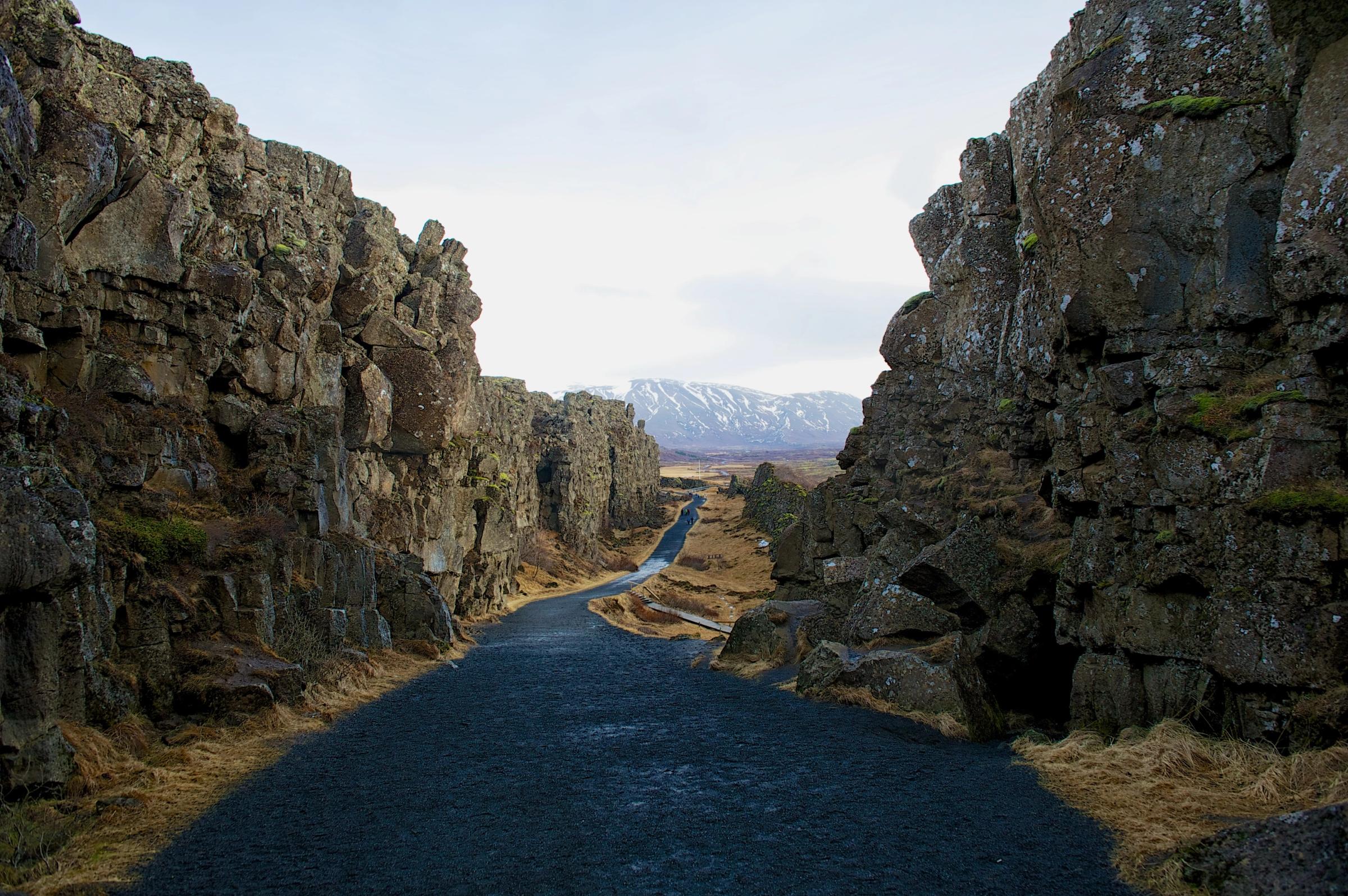 A dark path winds between two rugged rock walls toward distant snowy mountains under a cloudy sky.