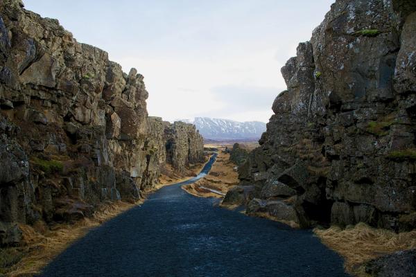 A dark path leads through a narrow gorge formed by towering rock walls, towards distant snow-capped mountains under a cloudy sky.