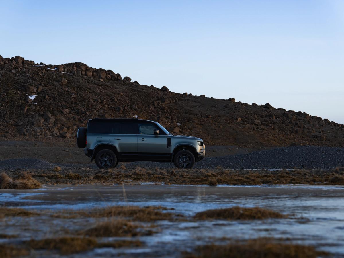 Vista lateral del Defender en un terreno rocoso Coche de alquiler Defender aparcado junto a un campo de lava rocosa en Islandia, disponible para alquilar en Go Car Rental