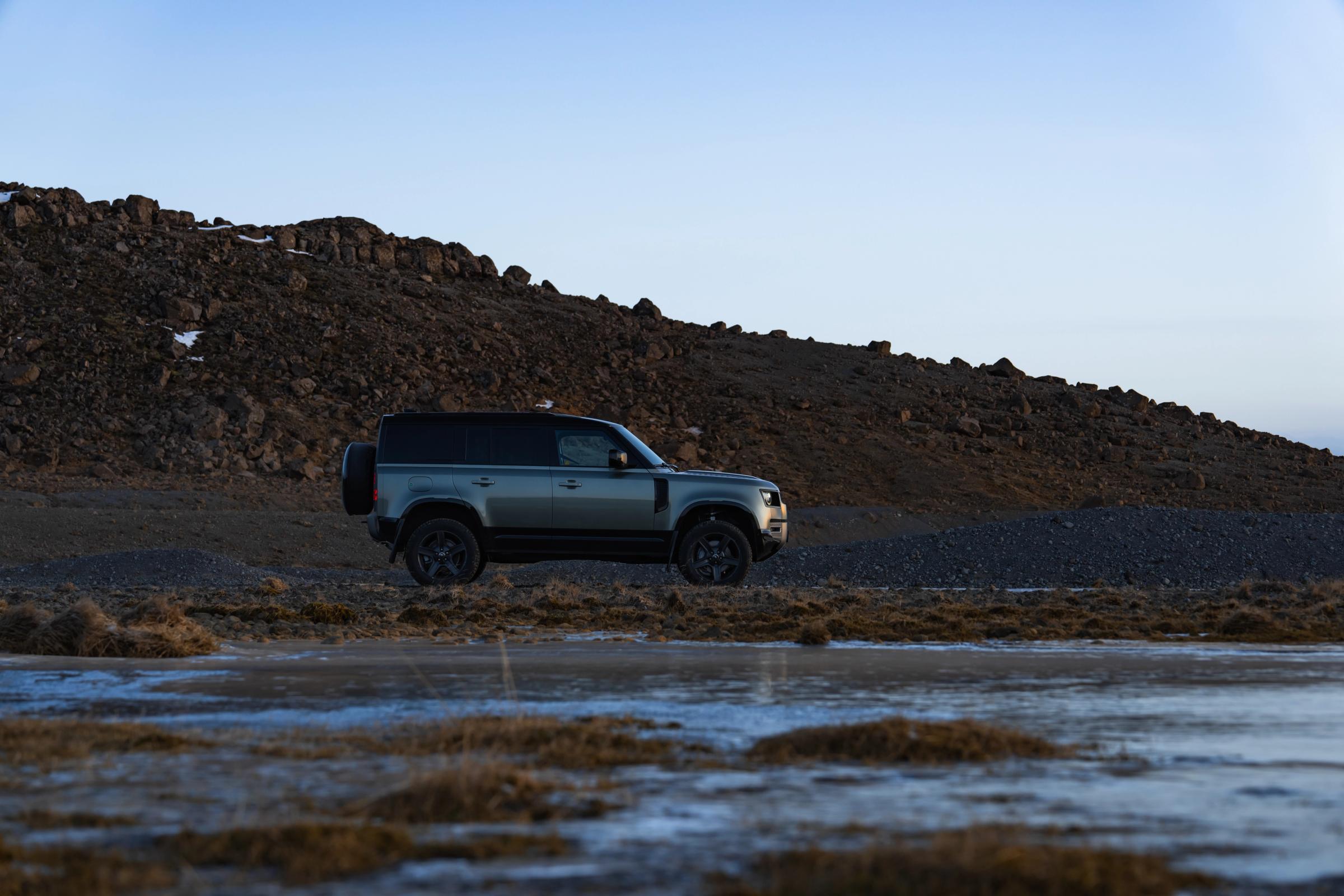 Vista lateral del Defender en un terreno rocoso Coche de alquiler Defender aparcado junto a un campo de lava rocosa en Islandia, disponible para alquilar en Go Car Rental