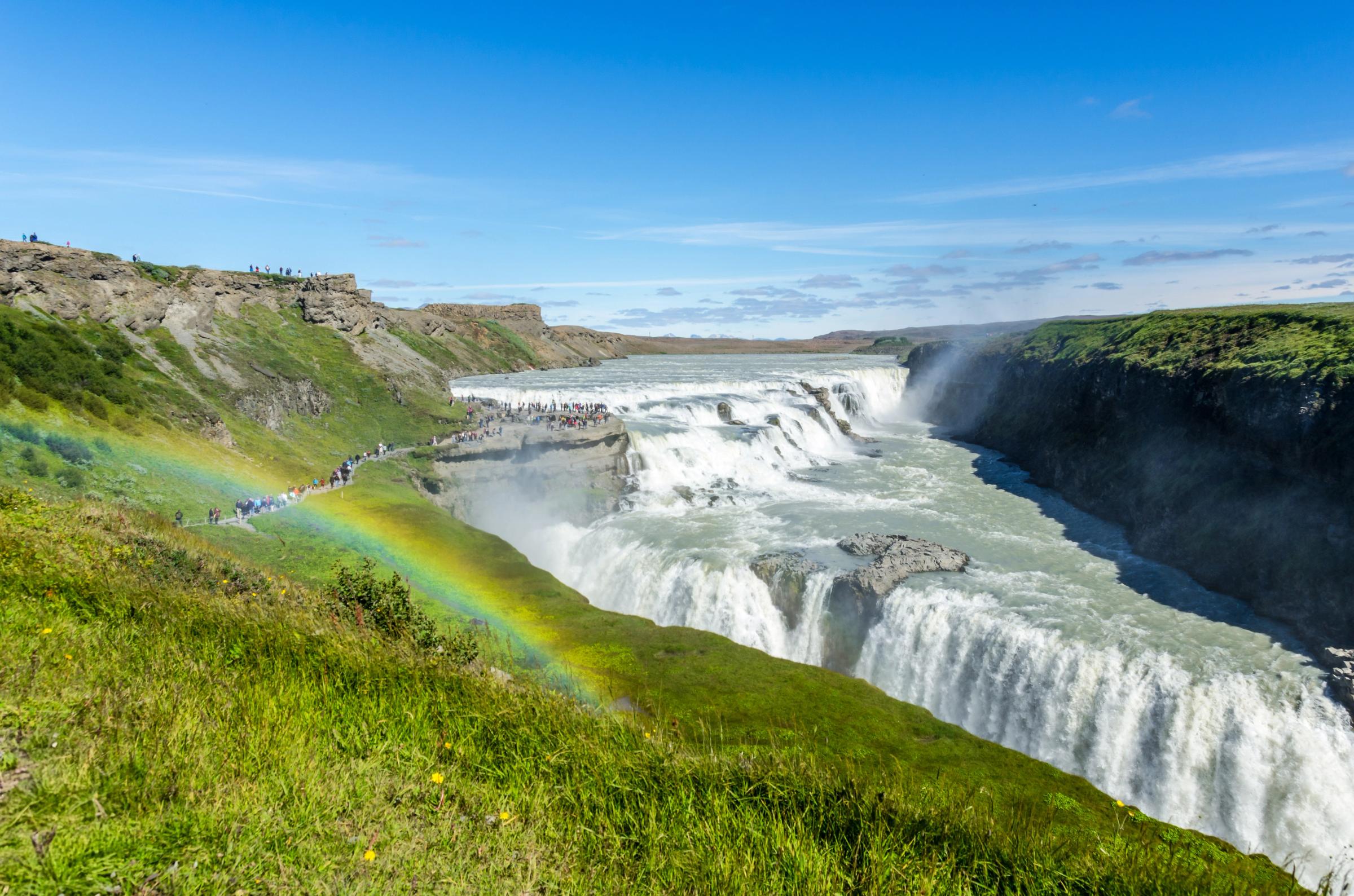 Una poderosa cascada de varios niveles cae en un cañón, con un vibrante arcoíris que se arquea sobre los acantilados cubiertos de hierba donde se reúnen los turistas bajo un cielo azul.