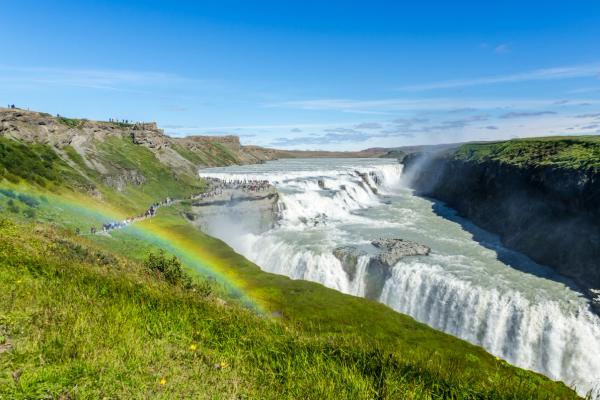 A large, multi-tiered waterfall with a rainbow in its mist, and many people on the green banks.