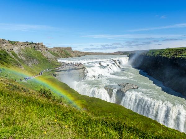 A powerful multi-tiered waterfall cascades into a canyon, with a vibrant rainbow arching over the grassy cliffs where tourists gather under a blue sky.
