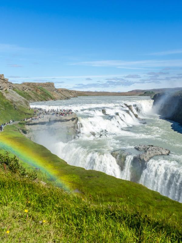 A powerful waterfall plunges into a canyon, with a vibrant rainbow arcing over green cliffs and many people observing.